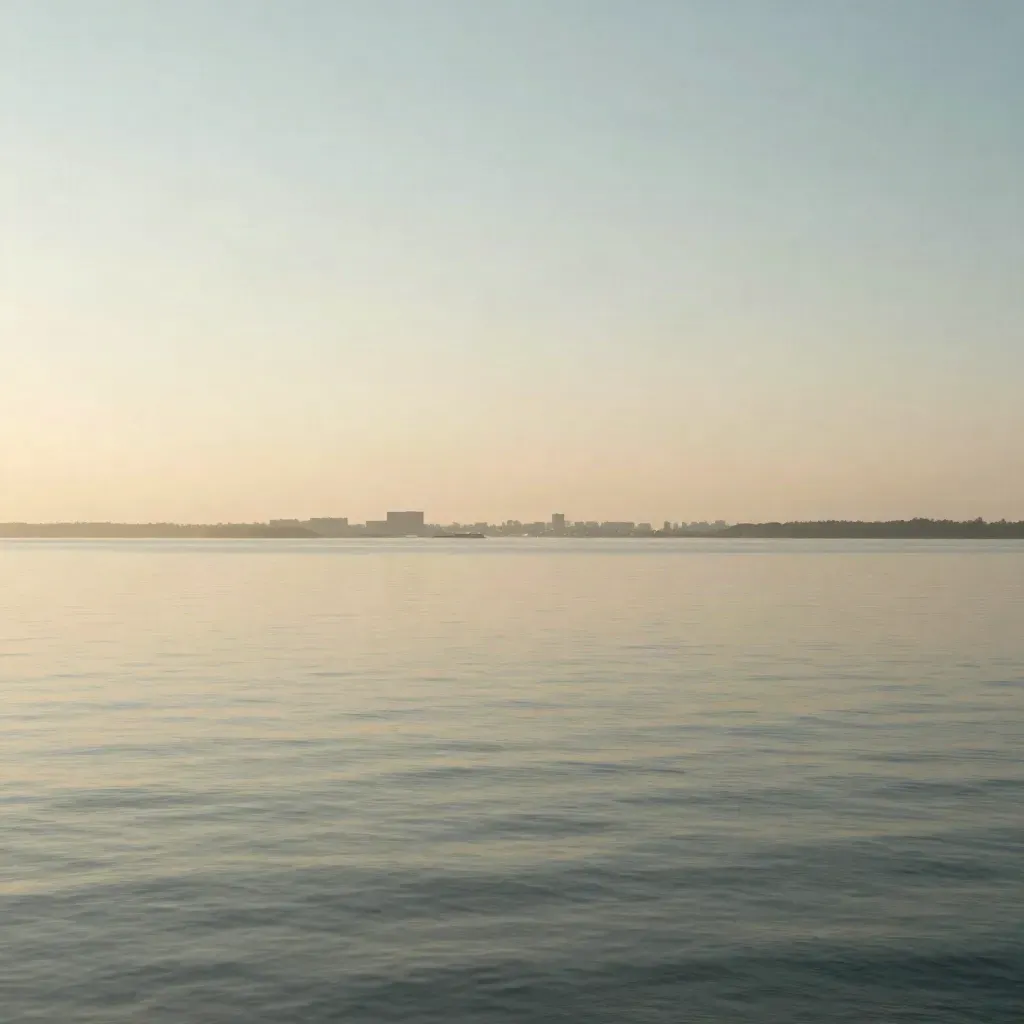 Hazy layered water, distant low city forms, and a calm sky in warm morning light.